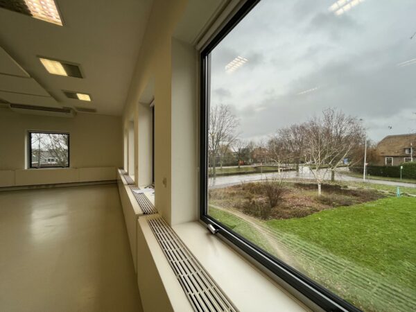 Uitzicht vanuit een leeg lokaal in Leidse Rijn op een nat en bewolkt straatbeeld met kale bomen en grasveld.